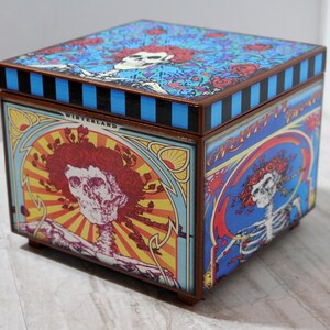 May include: A wooden box with a colorful design featuring a skull and flowers. The box has a blue and black striped lid and the text "Grateful Dead 1965-1970" on the side.