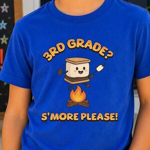 May include: A royal blue short-sleeved t-shirt featuring a graphic of a smiling s'more character above a campfire. The text above the graphic reads 3RD GRADE? and the text below reads S'MORE PLEASE! The shirt is made of a solid cotton fabric.