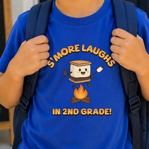 May include: A royal blue short-sleeved t-shirt featuring a graphic of a smiling s'more character jumping over a campfire. The text reads S'MORE LAUGHS IN 2ND GRADE! The shirt is worn with a dark blue backpack and light blue denim jeans.