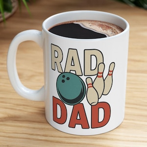 Peut inclure: Tasse à café en céramique blanche avec l'inscription "RAD DAD" dans une police rétro, accompagnée d'une boule de bowling turquoise et de quilles beiges. La tasse contient du café noir et est posée sur une surface en bois.