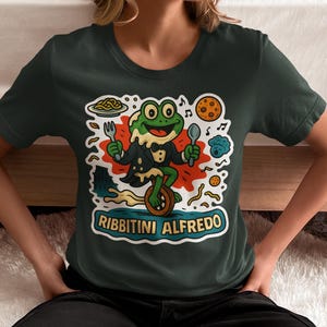 May include: Dark green t-shirt featuring a cartoon frog chef on a unicycle, holding a fork and spoon. The frog is wearing a tuxedo and the text "Ribbitini Alfredo" is displayed below the frog.