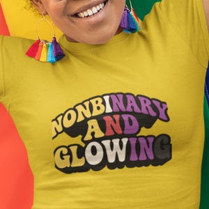 May include: Yellow t-shirt with the text "NONBINARY AND GLOWING" in a retro font. The shirt is a solid yellow color. The image also shows colorful earrings and a rainbow flag in the background.