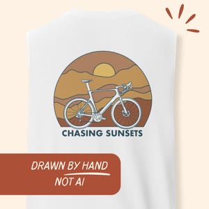 Puede incluir: Camiseta sin mangas blanca con un gráfico de bicicleta sobre un atardecer. El diseño incluye el texto "CHASING SUNSETS" y "DRAWN BY HAND NOT AI". El gráfico utiliza tonos marrones, naranjas y blancos.