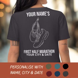 Op de afbeelding: Donkergrijs t-shirt met witte tekst en een lijntekening van hardloopschoenen. De tekst luidt "YOUR NAME'S FIRST HALF MARATHON YOUR CITY & DATE". De onderkant van het shirt heeft de tekst "PERSONALISE WITH NAME, CITY & DATE".