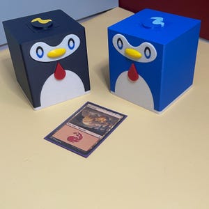 Könnte beinhalten: Zwei würfelförmige Aufbewahrungsboxen in Pinguinform, eine dunkelblau und eine hellblau, jeweils mit weißem Bauch, gelbem Schnabel, roter Fliege und weißen Augen mit blauen Pupillen. Eine einzelne Sammelkarte mit der Aufschrift Mountain liegt davor.