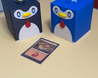May include: Two cubic storage boxes shaped like penguins, one dark blue and one bright blue, each featuring a white belly, yellow beak, red bowtie, and white eyes with blue pupils. A single trading card labeled Mountain is placed in front of them.