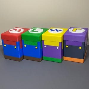 Puede incluir: Cuatro cajas de almacenamiento apiladas y coloridas, cada una con un tema de personaje diferente. Las cajas son rojas, verdes, amarillas y moradas, con detalles en azul y marrón. Cada caja tiene una tapa circular blanca con una inicial de personaje.