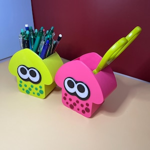 May include: Two squid-shaped desk organizers in bright lime green and hot pink. Each container features large white and black eyes and circular spots on the front. The lime green organizer holds various pens, while the pink one holds yellow scissors.