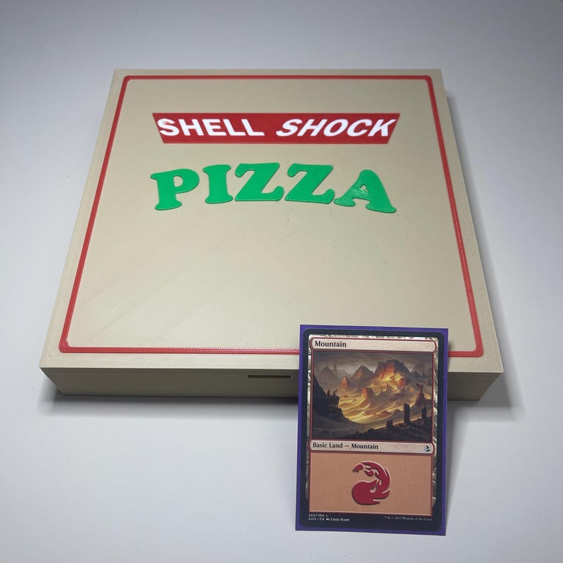 May include: A wooden pizza box with the text "SHELL SHOCK PIZZA" in red and green. A Magic: The Gathering card titled "Mountain" is in front of the box. The card has a purple border and features a landscape image.