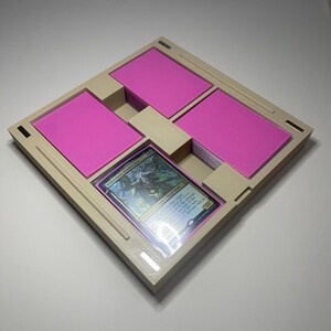 May include: A beige card organiser with four compartments, each holding stacks of pink cards. A single, holographic trading card is visible in a central compartment. The organiser is square with a textured surface.