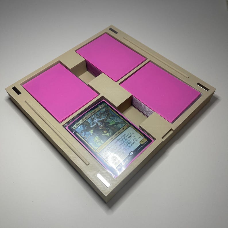 May include: A beige card organiser with four compartments, each holding stacks of pink cards. A single, holographic trading card is visible in a central compartment. The organiser is square with a textured surface.