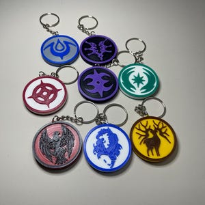 May include: A collection of round keychains with various colorful designs. Each keychain features a different emblem or symbol in colors like red, blue, purple, and yellow. The keychains are attached to silver key rings.