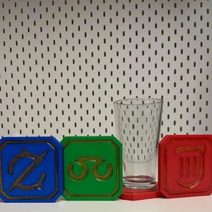 May include: A clear glass and four colorful game pieces. The game pieces are blue, green, and red, each with a unique gold symbol. The glass is tall and cylindrical, with a textured pattern.