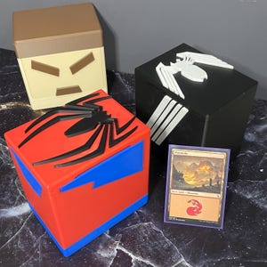 Puede incluir: Cuatro cajas de almacenamiento cúbicas con diseños de superhéroes. Una caja es roja y azul con un emblema de araña negra. Otra es negra con un emblema de araña blanca. Una tercera es beige y marrón. También se ve una carta.