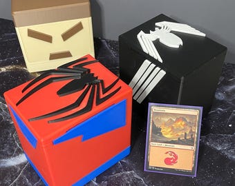 May include: Four cube-shaped storage boxes with superhero-themed designs. One box is red and blue with a black spider emblem. Another is black with a white spider emblem. A third is tan and brown. A card is also visible.