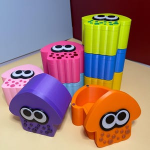May include: A collection of six plastic squid-shaped containers in various colours including lime green, pink, purple, and orange. Each container features large white and black eyes and small circular indentations on the front surface.
