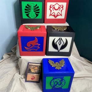 May include: A collection of colorful, cube-shaped storage boxes with gold-colored bird-shaped embellishments on top. Each box features a different color scheme and a unique, stylized graphic on the front. A trading card rests in front of the boxes.