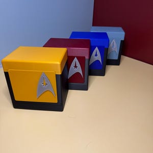 May include: Four square storage boxes with black bases and colored lids in yellow, red, blue, and light blue. Each box features a silver Starfleet delta insignia emblem centered on the front panel, arranged in a diagonal line on a flat surface.