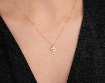 May include: A delicate gold necklace featuring a sunburst pendant with a small, dark center stone. The pendant is suspended from a fine gold chain. The necklace is shown against a dark background.