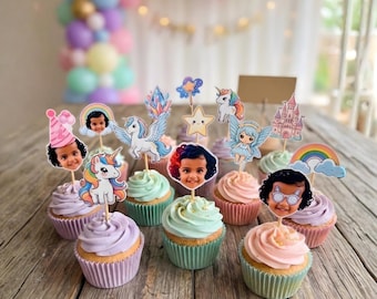 May include: A collection of cupcakes with pastel frosting, each topped with a decorative pick featuring a child's face and whimsical designs like unicorns, rainbows, and a castle. The cupcakes sit on a wooden surface.