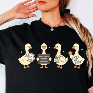 May include: A black t-shirt featuring a graphic of four light yellow ducks. The first holds a clipboard, the second a sign reading ABCs of Behavior Antecedent Behavior Consequence, the third wears a lanyard, and the fourth holds a star and paper.