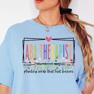 Op de afbeelding: Een lichtblauw t-shirt met een afbeelding met de tekst ABA THERAPIST in veelkleurige letters, een klein rood hartje en een bloemenrand. Onder de rand staat de tekst planting seeds that last forever in een zwart cursief lettertype.