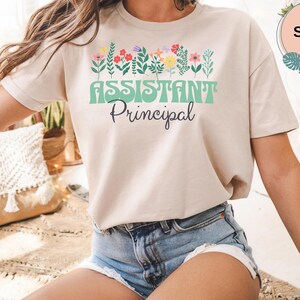Op de afbeelding: Een zandkleurig t-shirt met een afbeelding van kleurrijke wilde bloemen in roze, gele en groene tinten. Onder de bloemen staat de tekst "ASSISTANT" in een groot, lichtgroen retro lettertype, gevolgd door het woord "Principal" in zwart schrift.