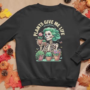 May include: Black sweatshirt with a colourful illustration of a skeleton with green hair holding a potted plant. The text "PLANTS GIVE ME LIFE" is arched above the skeleton. The design includes butterflies and other potted plants.