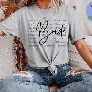 Könnte beinhalten: Ein sportgraues Kurzarm-T-Shirt mit schwarzem Text, der Adjektive wie Radiant, Elegant, Beautiful, Joyful, Glamorous, Charming, Alluring, Refined, Graceful, Breathtaking, Classic, Romantic, Blissful, Vibrant, Lovely, Poised, Loyal, Chic, Timeless, Stunning, Glowing, Exquisite, Delicate, Serene auflistet, sowie das Wort Bride in einer großen schwarzen Schreibschrift in der Mitte.