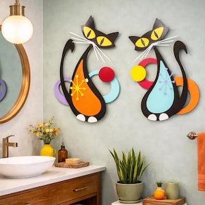 May include: Two wall-mounted cat decorations featuring black silhouettes with yellow eyes and white whiskers. The left cat has an orange body with a yellow starburst, while the right cat has a light blue body with a light blue starburst, both accented by colorful geometric circles.
