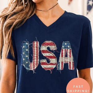 Könnte beinhalten: Marineblaues V-Ausschnitt-T-Shirt mit dem Wort "USA" in einem verwitterten Design der amerikanischen Flagge. Die Buchstaben sind rot, weiß und blau. Das Model trägt Jeansshorts. Das Bild hat ein patriotisches Thema.