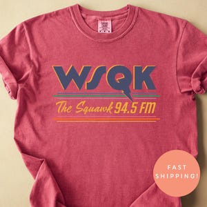 May include: Crimson-colored t-shirt with the text "WSQK The Squawk 94.5 FM" in navy blue, green, and orange. The shirt has rolled-up sleeves and a round neck. A peach-colored circle with the words "Fast Shipping!" is on the right side.