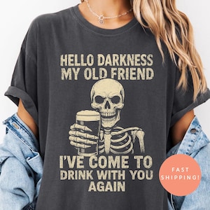 K&ouml;nnte beinhalten: Dunkelgraues T-Shirt mit einem Skelett, das ein Bier h&auml;lt, und dem Text "HELLO DARKNESS MY OLD FRIEND I'VE COME TO DRINK WITH YOU AGAIN." Das Shirt hat eine lockere Passform.