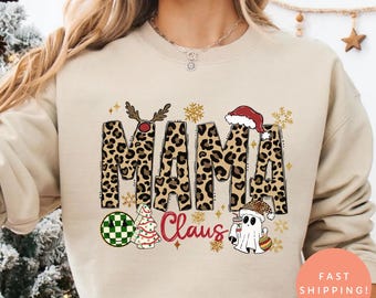 May include: A beige sweatshirt with the words "MAMA Claus" in a leopard print design. The design includes Christmas-themed graphics like a reindeer, Santa hat, and a ghost. The sweatshirt is paired with red and white plaid pajama pants.