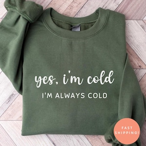 Może przedstawiać: Zielona bluza w kolorze oliwkowym z napisem "yes, i'm cold" i "I'M ALWAYS COLD" w białym skrypcie. Bluza jest starannie złożona, prezentując okrągły dekolt i długie rękawy. Tło to jasny wzór drewna.