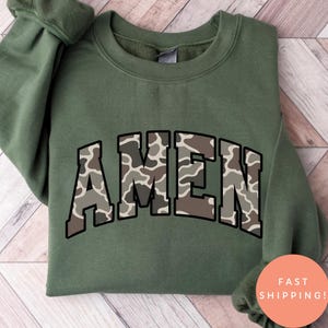 May include: An olive green sweatshirt with the word "AMEN" in a camouflage pattern across the chest. The sweatshirt has a crew neck and long sleeves. The text is outlined in a dark brown color.
