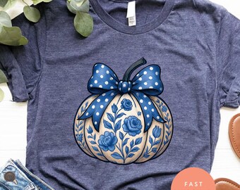 Könnte beinhalten: Marineblaues T-Shirt mit einem dekorativen Kürbis-Design. Der Kürbis ist mit blauen Blumenmustern und einer gepunkteten Schleife verziert. Die Abbildung zeigt auch Jeansshorts und Sandalen, was einen lässigen, herbstlichen Look suggeriert.