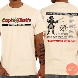 Peut inclure: Un t-shirt couleur cr&egrave;me avec un graphisme sur le devant indiquant Cap'n Clark's Ottoman Empire. Le dos pr&eacute;sente une illustration de pirate, une barre de navire, une liste de meubles, le texte 408-357-2875 et Everything Must Go.