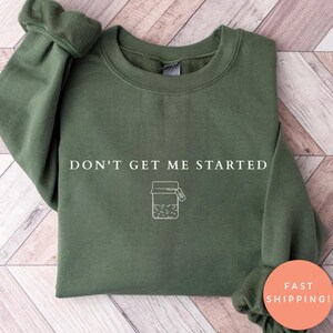 May include: An olive green sweatshirt with the phrase "DON'T GET ME STARTED" printed in white. Below the text is a white graphic of a jar with a tag. The sweatshirt has long sleeves and a crew neck.