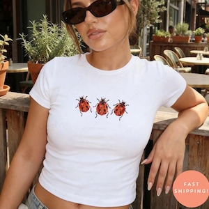 May include: A white short-sleeved t-shirt featuring a graphic print of three red ladybugs with black spots arranged in a horizontal row across the center of the chest. The shirt has a crew neckline and a fitted silhouette made of cotton fabric.