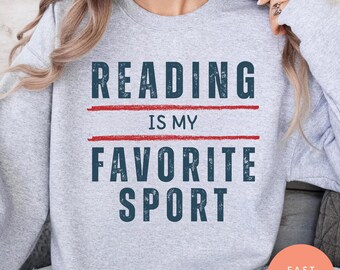 May include: A light gray sweatshirt with the text "READING IS MY FAVORITE SPORT" in navy blue letters, with a red line above and below the words "IS MY". The sweatshirt has long sleeves.