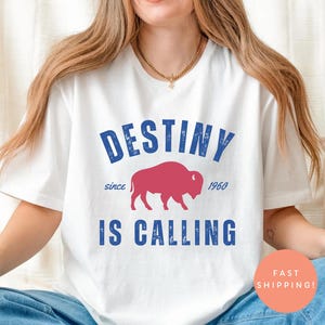 May include: White t-shirt with the text "DESTINY IS CALLING" in blue, featuring a red bison graphic. The shirt includes the text "since 1960". The shirt is a casual wear item.