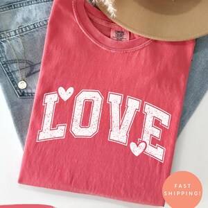 May include: A watermelon-colored t-shirt with the word "LOVE" in white, collegiate-style lettering. White hearts accent the letters. The shirt is folded on top of blue jeans, with a tan hat and necklace visible.