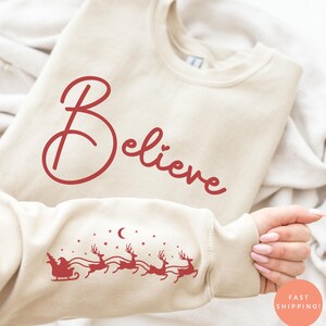 Könnte beinhalten: Cremefarbenes Sweatshirt mit dem Wort "Believe" in roter Schrift. Der Ärmel zeigt eine rote Grafik von Santa Claus in einem Schlitten, der von Rentieren unter einem Mond und Sternen gezogen wird. Ein runder Aufkleber mit der Aufschrift "Fast Shipping!" ist sichtbar.
