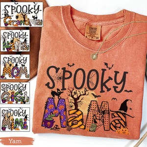 May include: A yam-colored Comfort Colors t-shirt with a round neck and a gold necklace. The shirt features the word "Spooky" in black, with "Mama" in orange and purple Halloween-themed letters. Bats, a witch hat, and pumpkins are also visible.