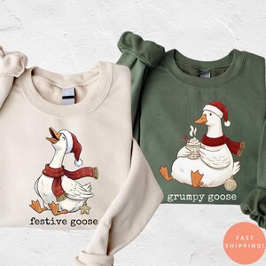 May include: Two sweatshirts, one cream and one green, with goose illustrations. The cream sweatshirt shows a goose in a Santa hat and scarf, with "festive goose" text. The green sweatshirt has a goose with a Santa hat, scarf, and mug, with "grumpy goose" text.