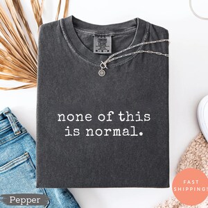 May include: A dark gray t-shirt with the phrase "none of this is normal" in white text. The shirt is folded and accessorized with a silver necklace. A pair of blue jeans and a dried palm leaf are also in the image.
