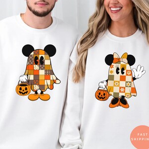 May include: White sweatshirts featuring Halloween-themed cartoon characters. One sweatshirt has a ghost-like character with a patchwork design, holding a pumpkin bucket. The other sweatshirt has a similar character with a bow and flowers, also holding a pumpkin bucket.