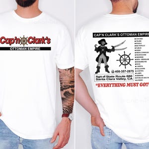 Peut inclure: Un t-shirt blanc &agrave; manches courtes avec un graphisme frontal portant le texte rouge Cap'n Clark's et une barre de navire. Le dos pr&eacute;sente une illustration de pirate, le texte Cap'n Clark's Ottoman Empire, une liste de meubles et Everything Must Go.