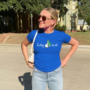 May include: A bright blue t-shirt with the words "lucky duck" and a duck graphic with a shamrock. The shirt is worn with light blue jeans. A white shoulder bag is visible. The image is taken outdoors.